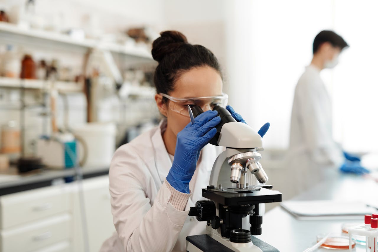 A female scientist in protective gear examines samples through a microscope in a laboratory setting.