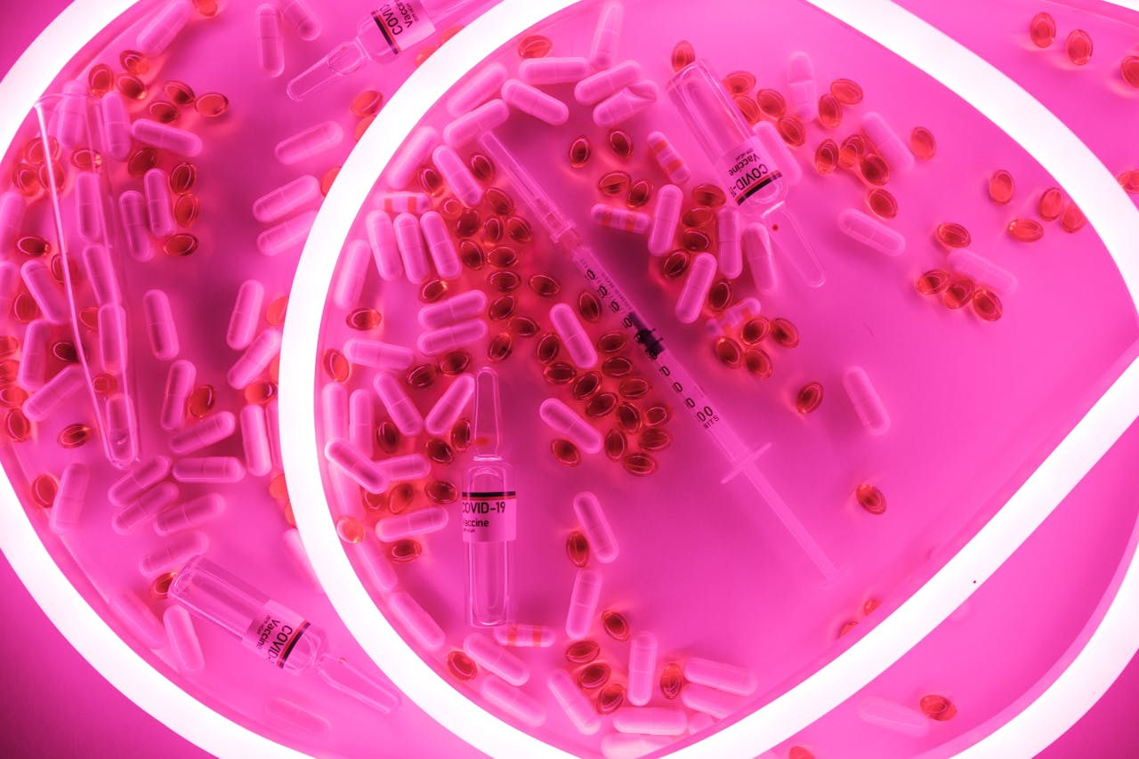 A vibrant neon pink composition of Covid-19 vaccine vials, capsules, and syringe viewed from above.